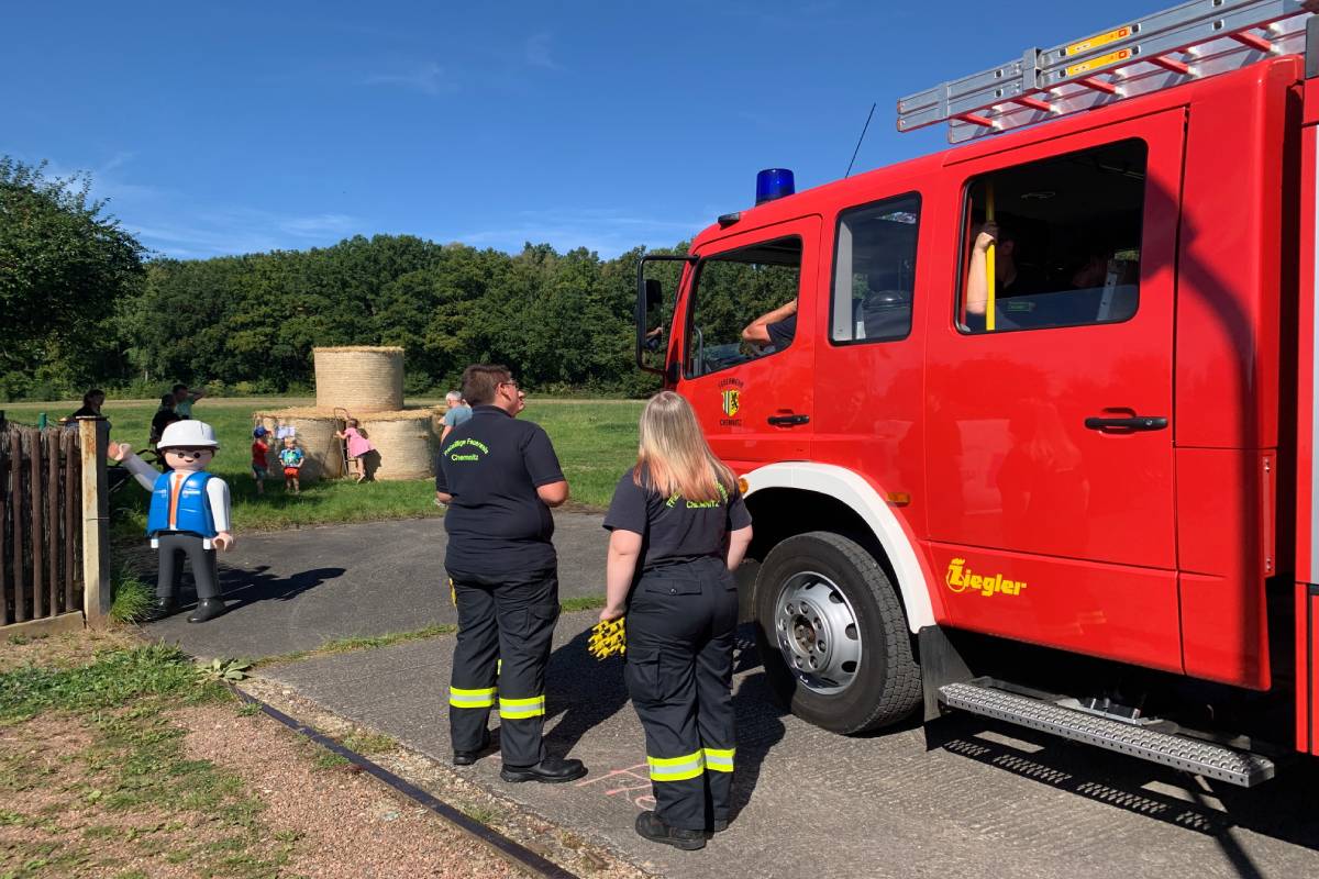 Zwei Feuerwehrleute vor einem Feuerwehrauto. Im Hintergrund spielende Kinder