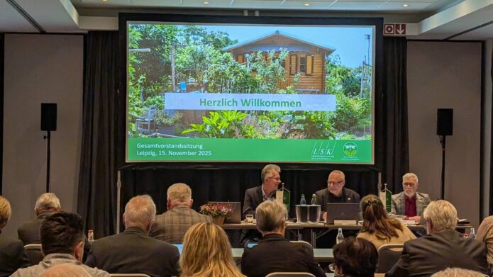 Blick auf die Präsentation auf der Leinwand mit dem Schriftzug "Herzlich Willkommen". Davor die Mitglieder des Präsdiums sowie ein Teil der Gäste
