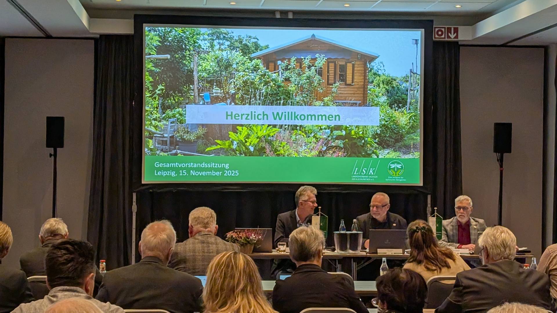 Blick auf die Präsentation auf der Leinwand mit dem Schriftzug "Herzlich Willkommen". Davor die Mitglieder des Präsdiums sowie ein Teil der Gäste